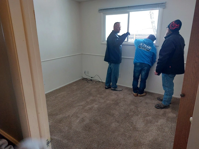 Three men are discussing a window in a sparsely furnished room with beige carpet and light-colored walls.